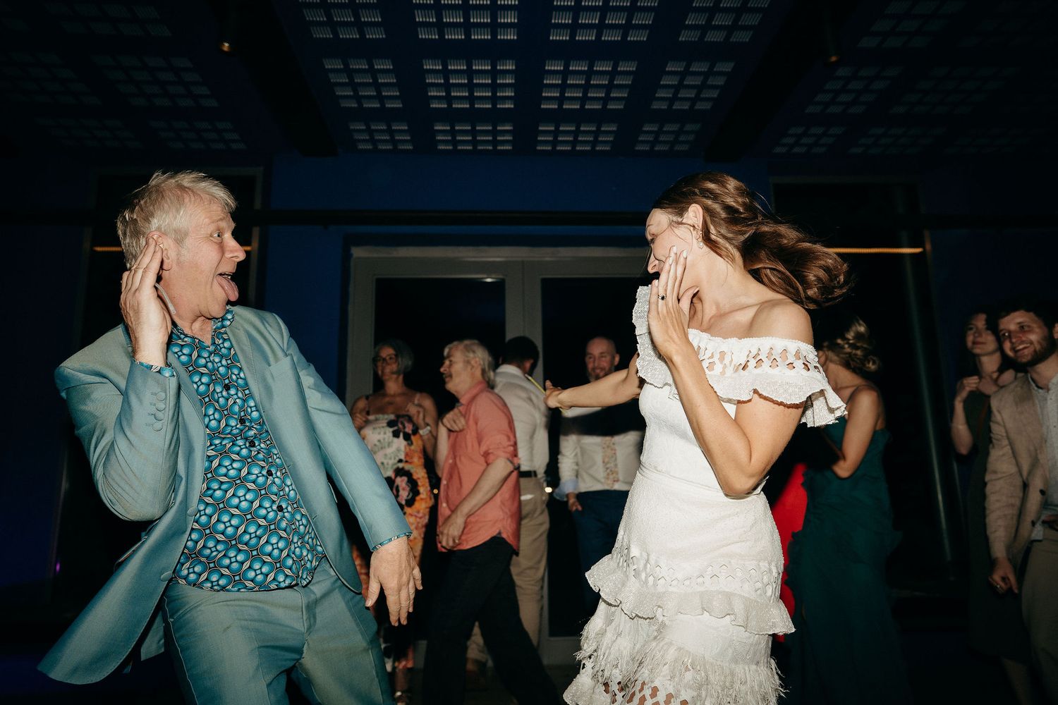 A bride in a white ruffled dress dances energetically with a guest during a lively reception party.