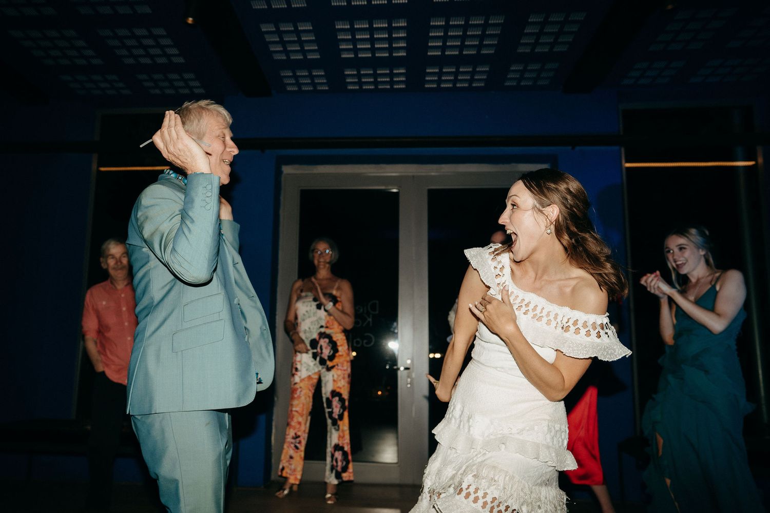 Two people dancing energetically on a dance floor during a celebration with guests watching in background.