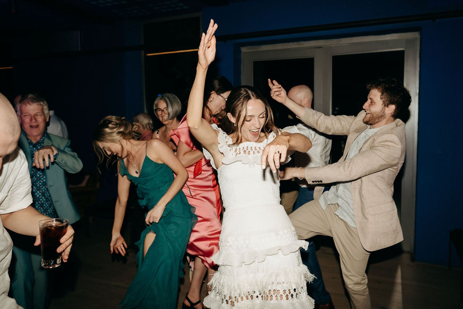 Wedding guests dancing energetically at a reception with people in green dresses and formal attire on the dance floor.