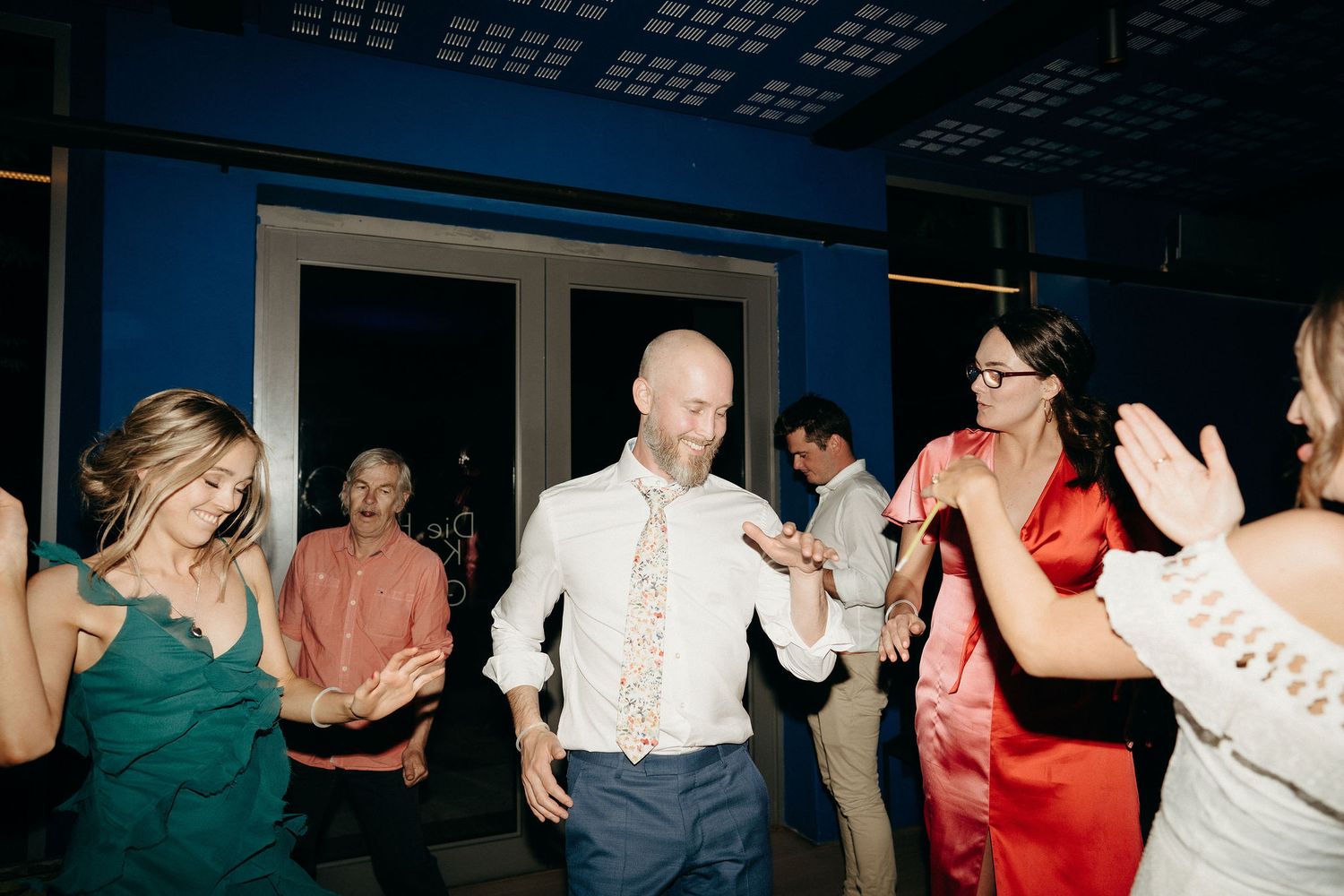 Partygoers dance and celebrate on the dance floor at an indoor venue with dark blue lighting and festive atmosphere.