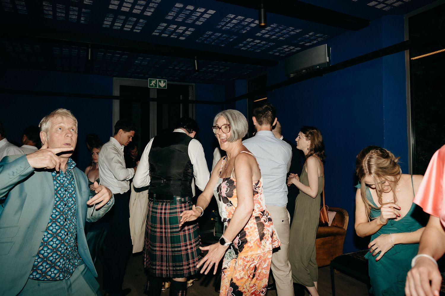 People dance and socialize at a dimly lit party venue with blue walls.