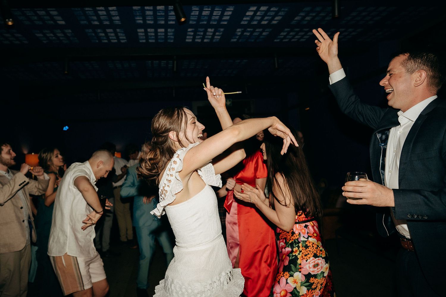 Wedding reception guests dancing and celebrating on a dark dance floor with arms raised.