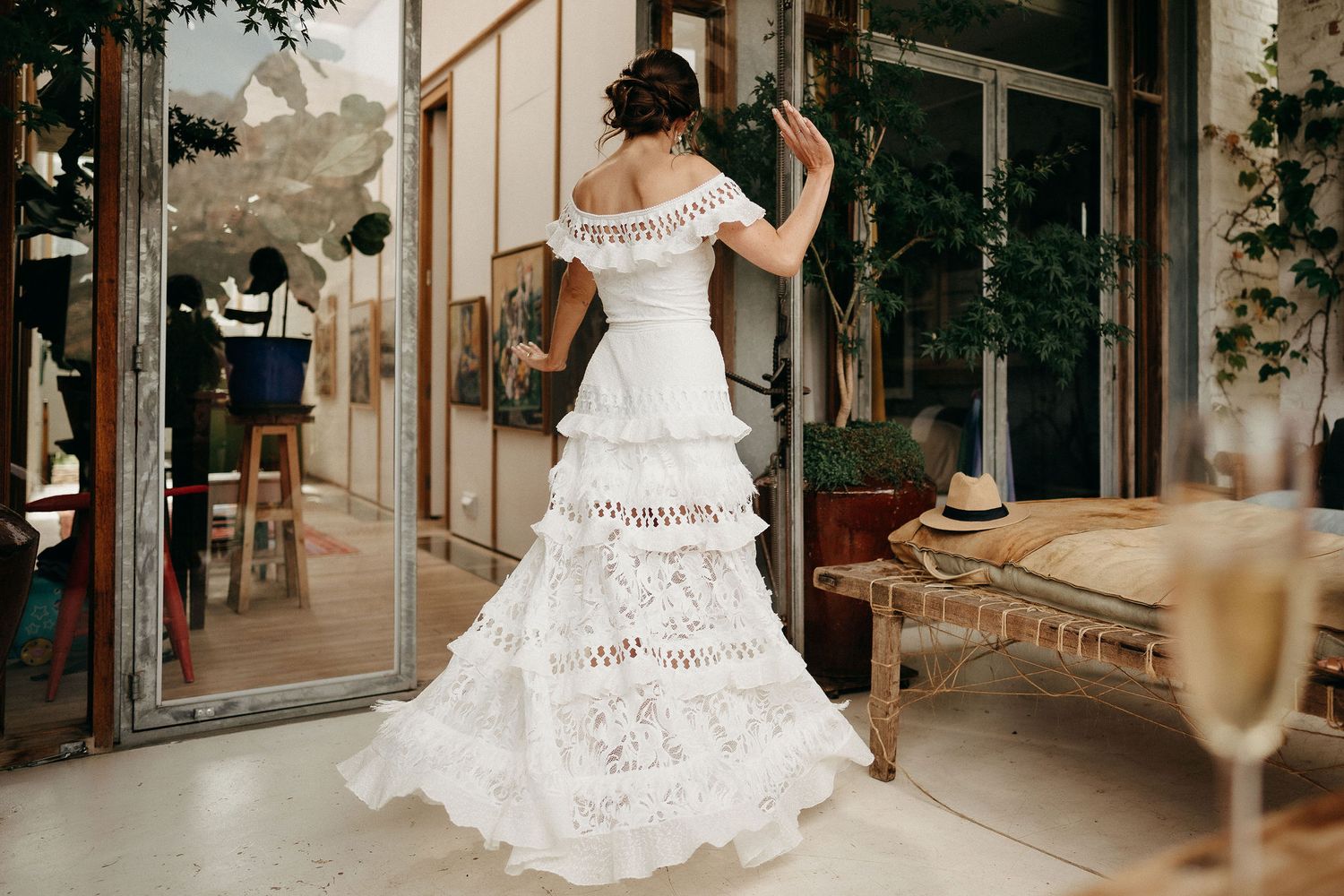 A white lace wedding dress with tiered ruffles is displayed on a figure standing near glass doors on a patio.