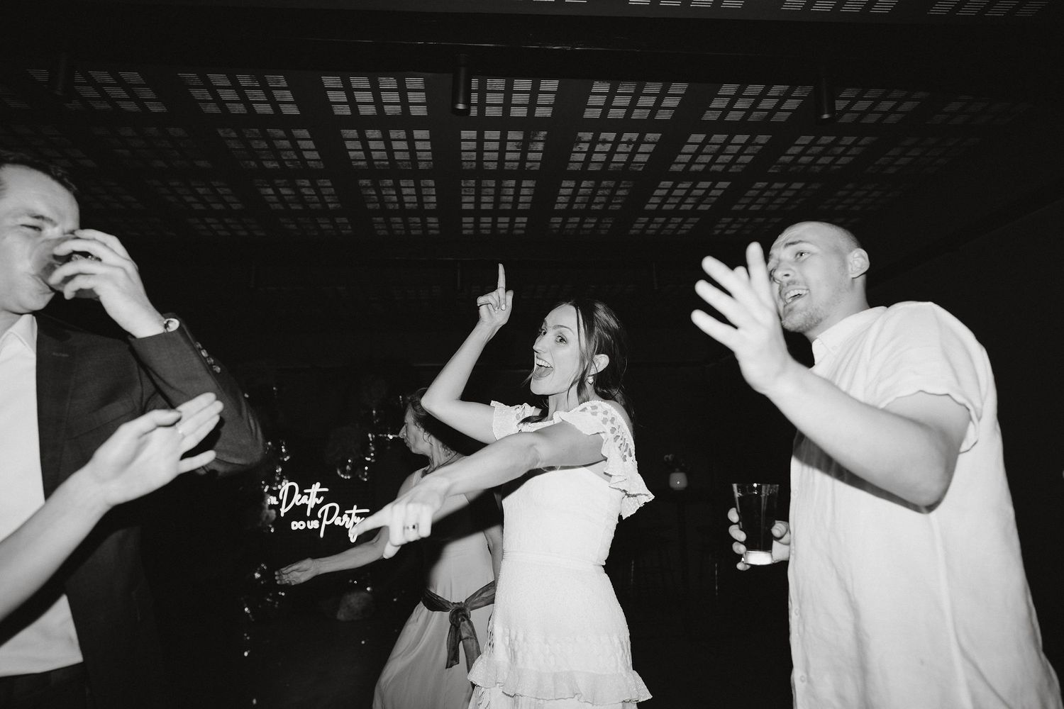 People dance enthusiastically at a party under an industrial-style ceiling in black and white photography.