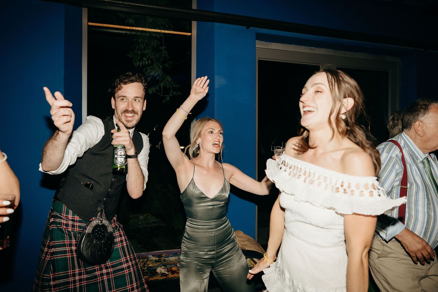 Wedding guests dancing and celebrating at a reception with someone wearing a traditional kilt among the crowd.