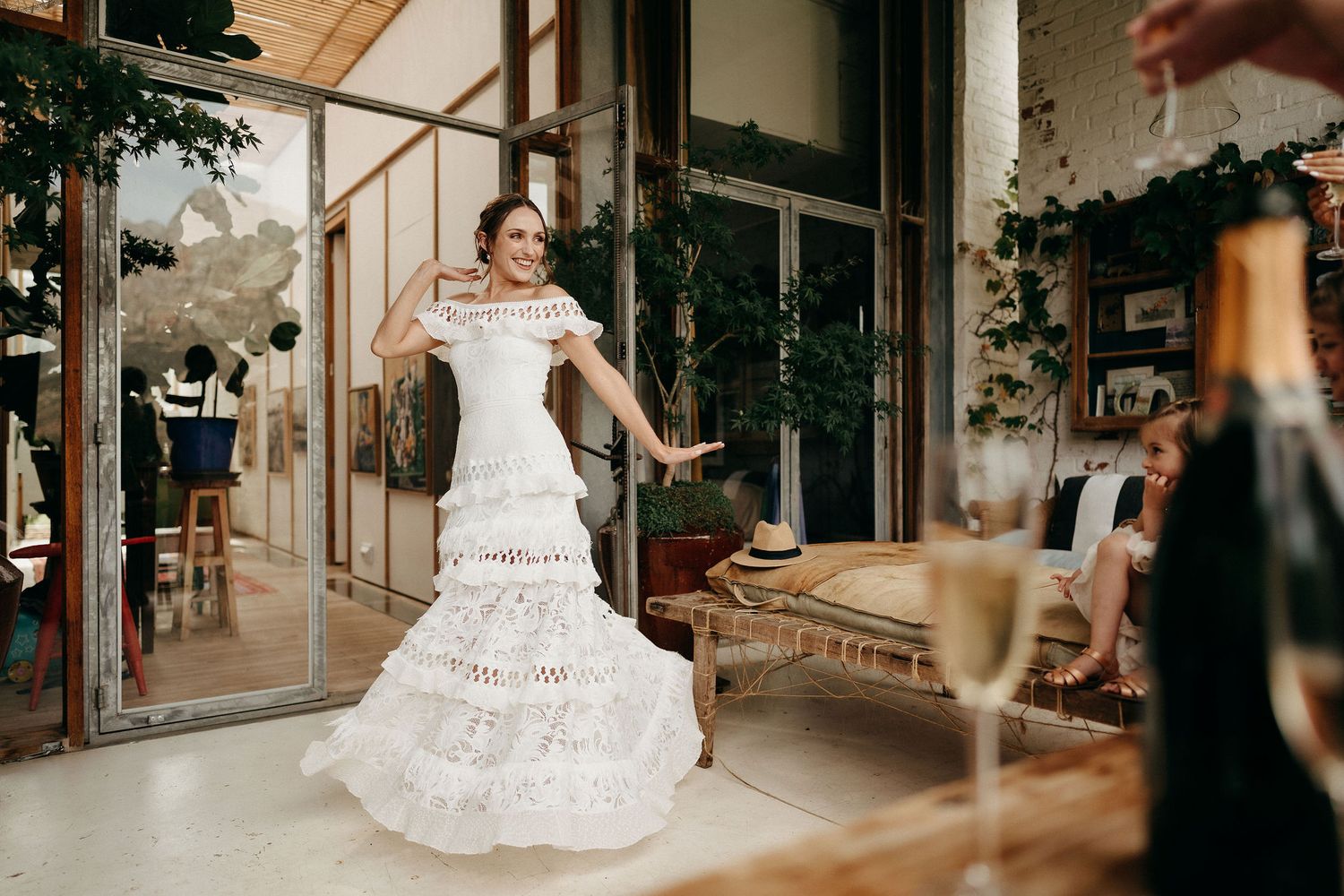 A bride in an elegant off-shoulder white ruffled wedding gown poses gracefully in front of a rustic venue entrance.