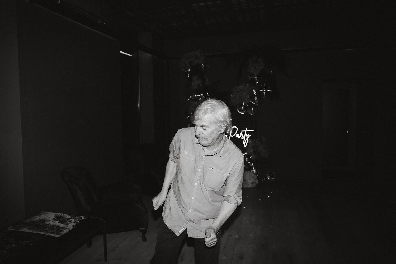 A black and white photograph shows someone dancing in a dark room with a drum kit visible in the background.