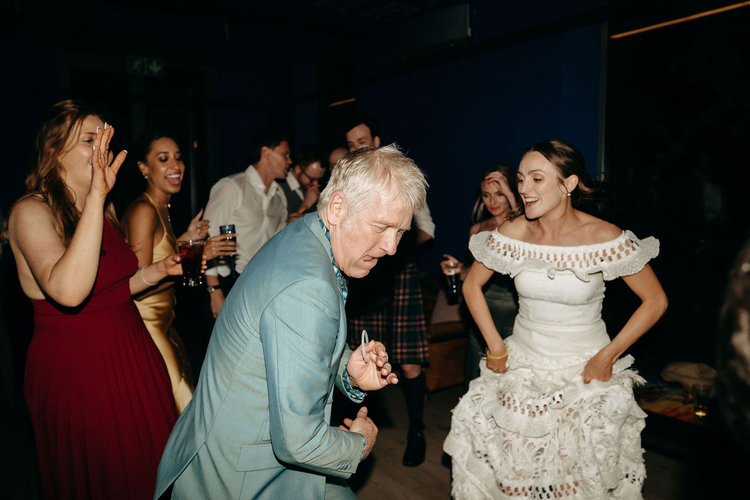 Party guests dance and celebrate at a wedding reception with a person in a ruffle white dress and others in formal attire.