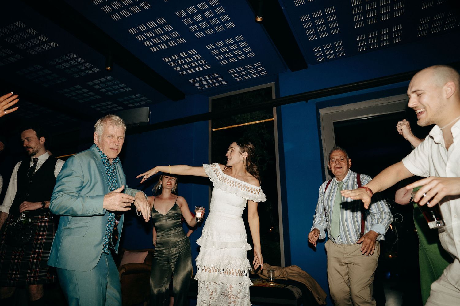 Wedding guests dancing together and having fun on a dance floor at an indoor reception with blue lighting overhead.