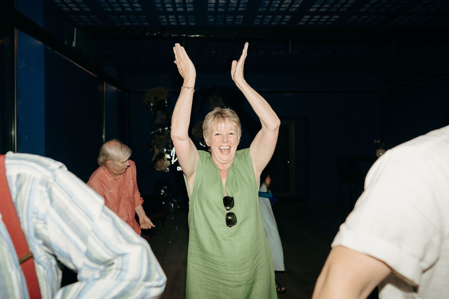 A person in a sage green outfit dances with raised arms in a dimly lit room with other partygoers.