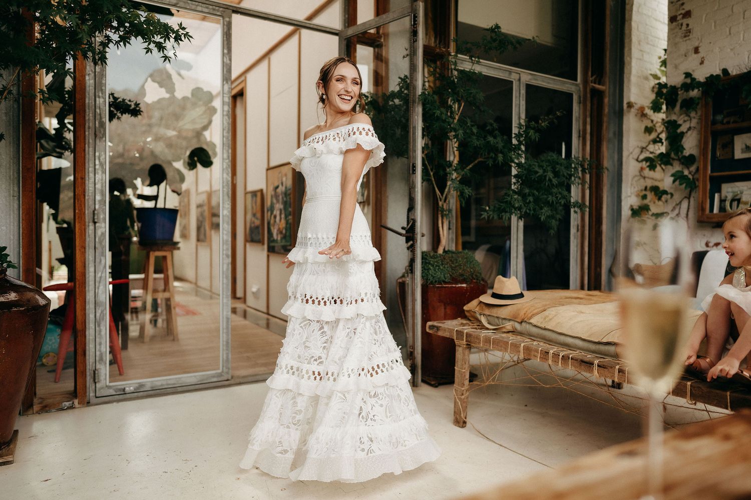Elegant lace wedding dress with tiered skirt and cap sleeves photographed in a sunlit greenhouse setting.