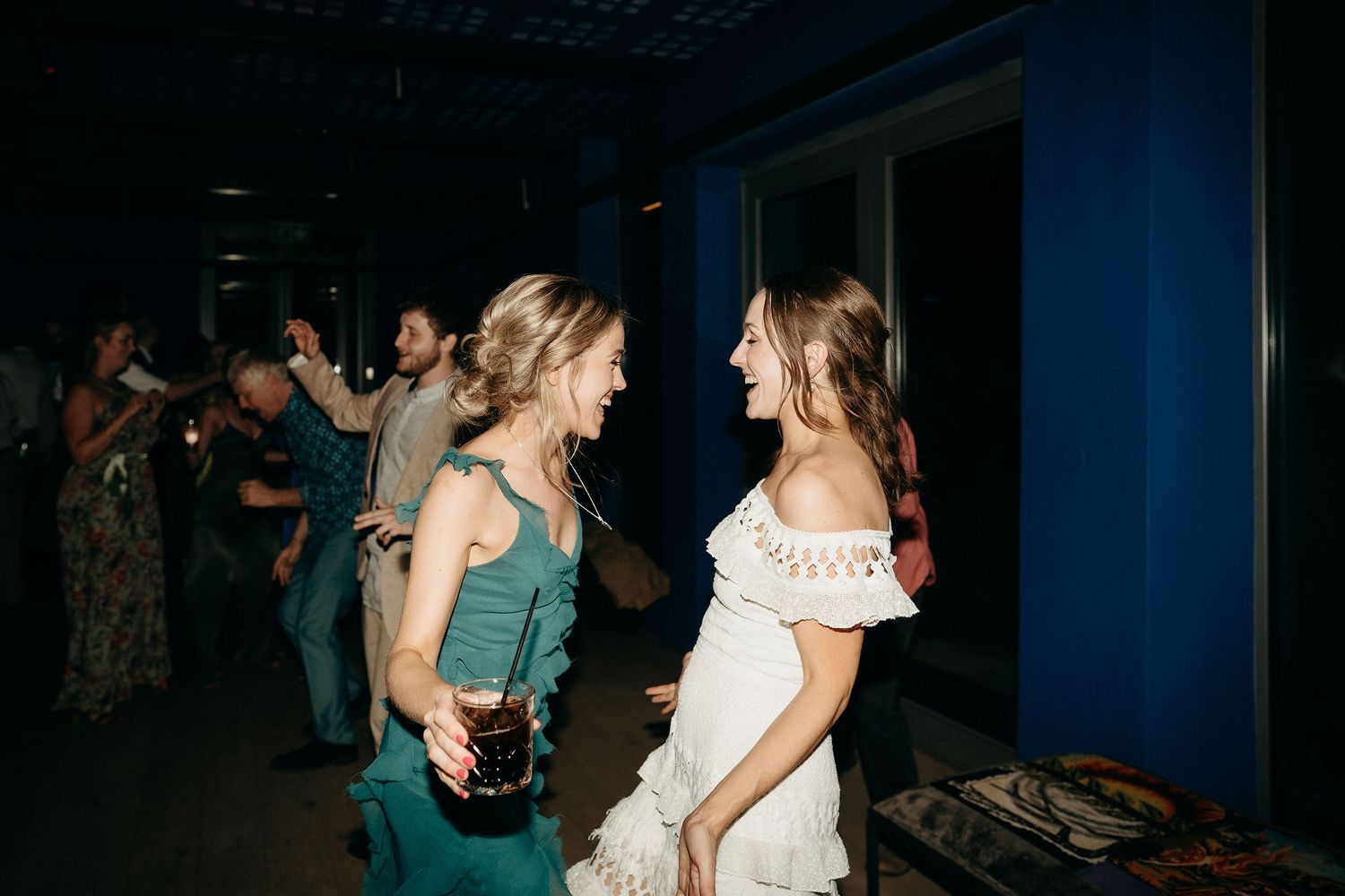 Two women in casual evening wear dance and smile together at a dimly lit indoor venue.