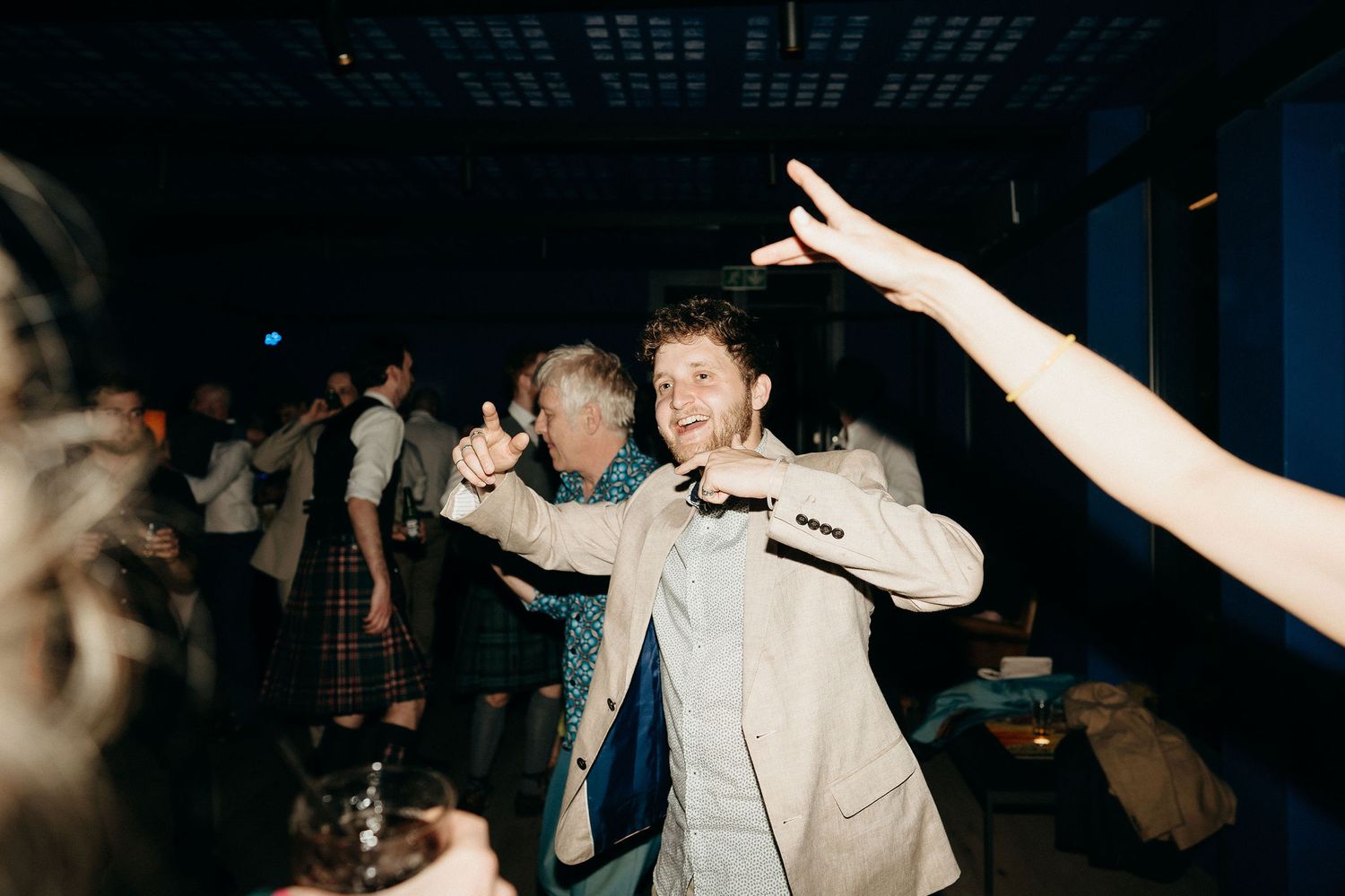 People dancing and socializing at a dimly lit party or nightclub with raised hands and energetic movement in view.