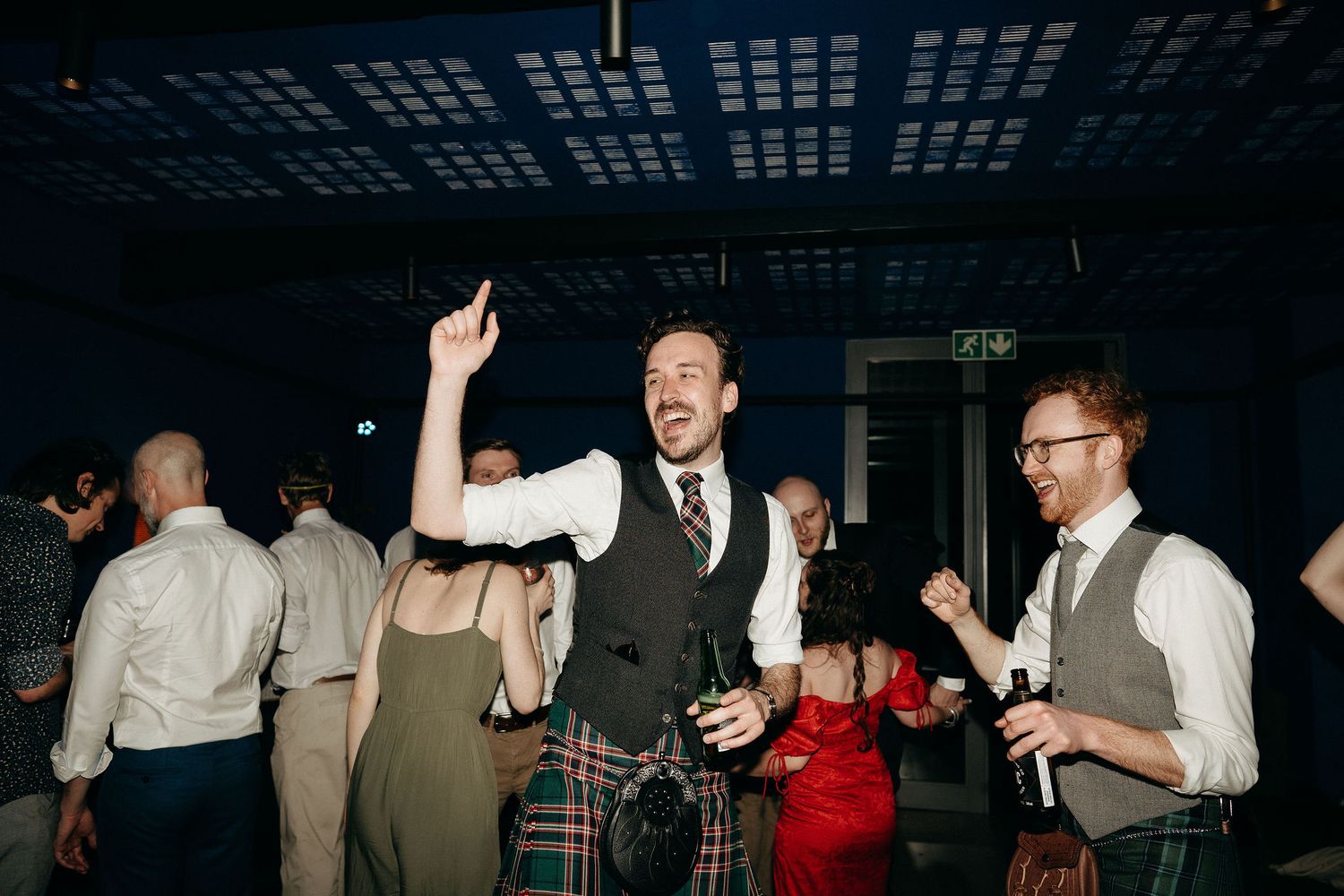 Wedding guests dancing and celebrating at an indoor reception with dark ambient lighting.