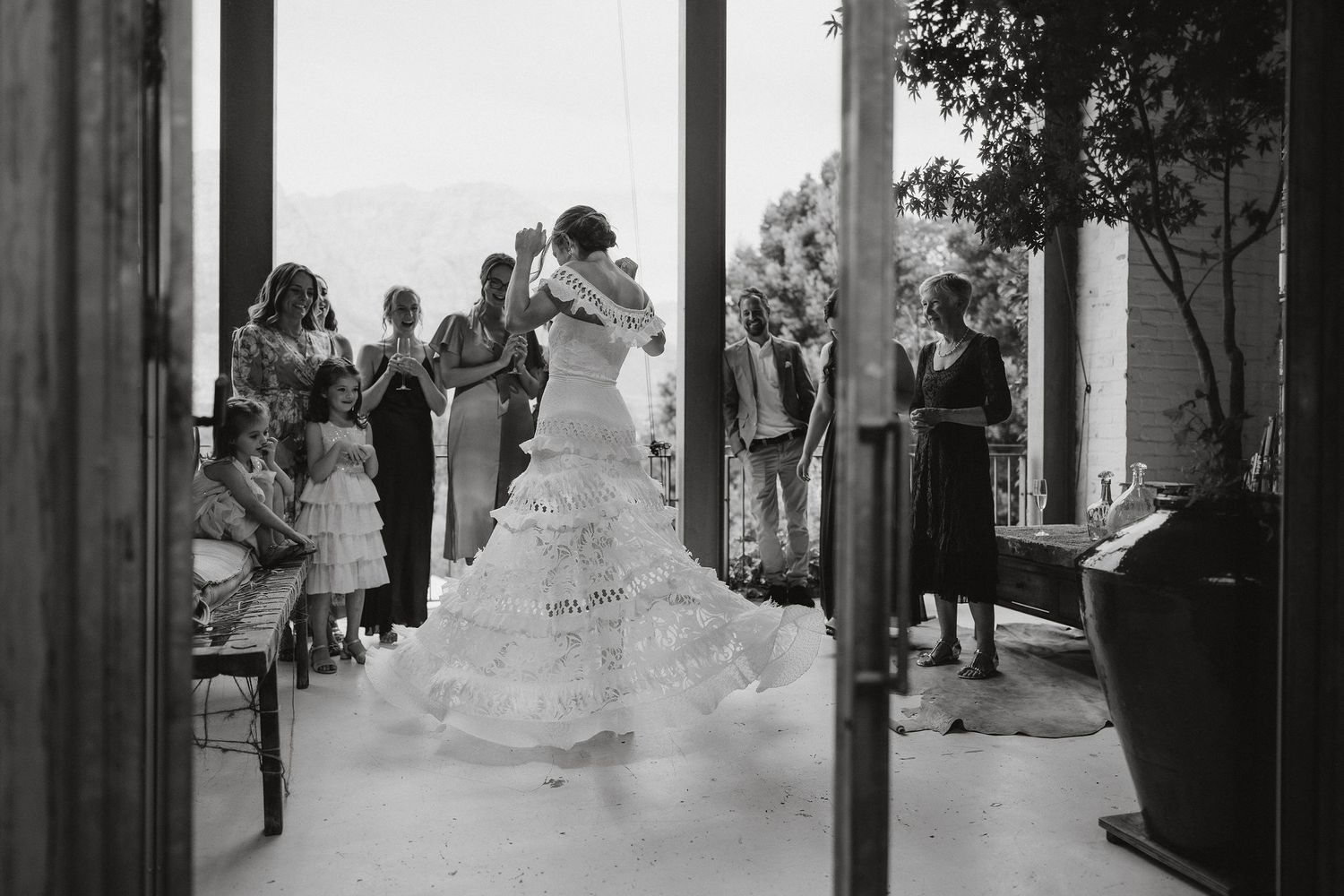 Black and white photo capturing a wedding ceremony moment with the bride in a ruffled dress standing among guests.