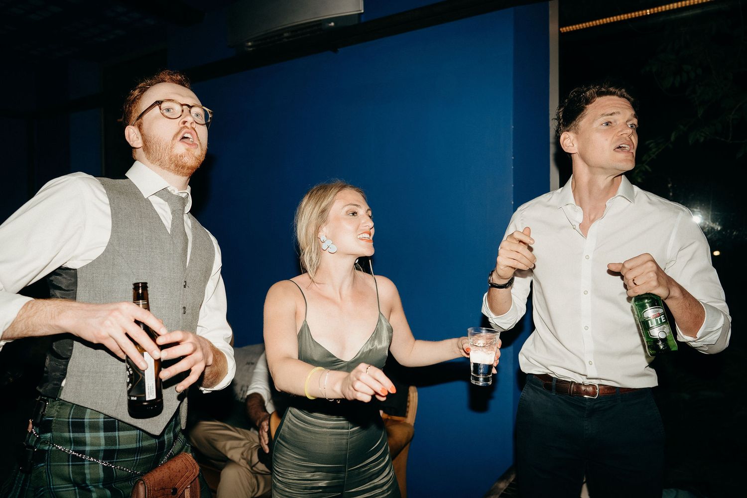 Party guests in casual attire laugh while holding drinks at a social gathering against a blue wall.