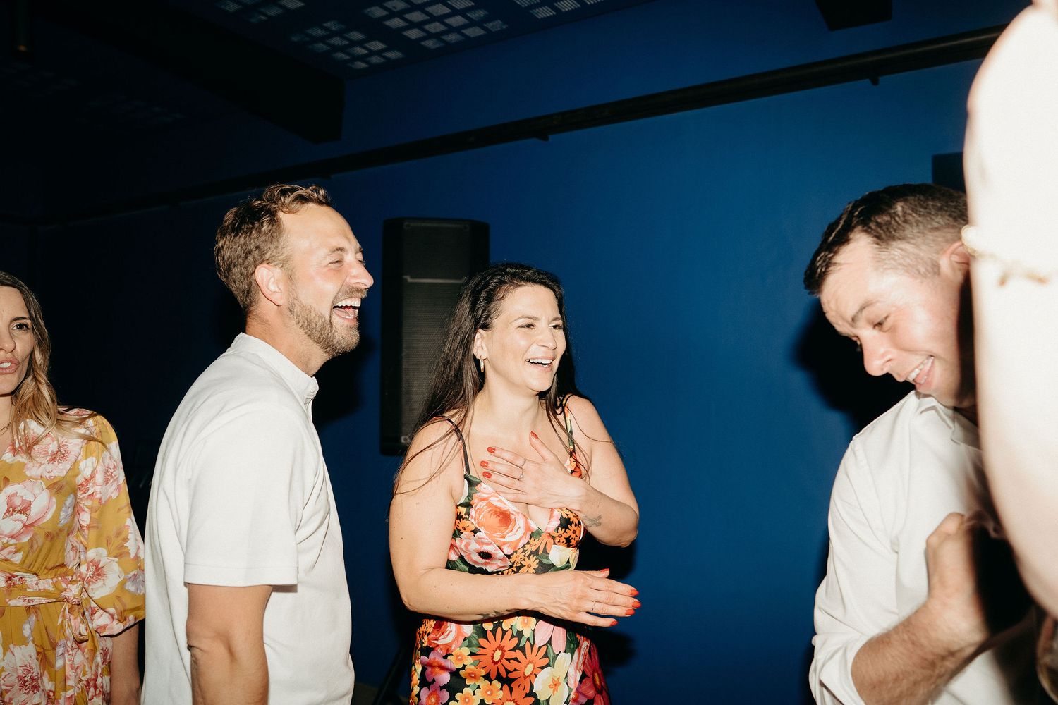 A group of people laugh together at a casual social gathering in a dark room with blue lighting.