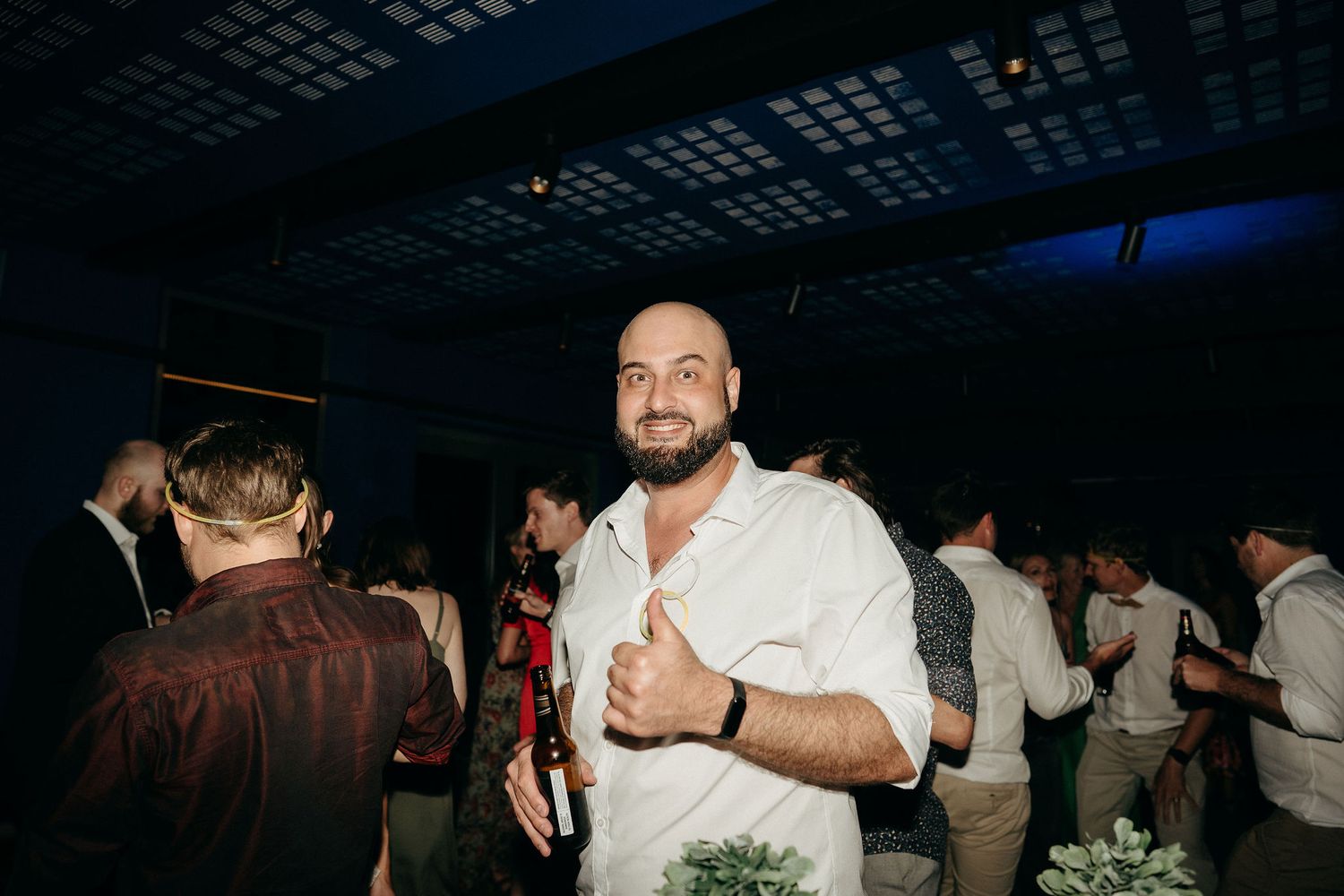 A person in a white shirt gives a thumbs up while smiling at a dimly lit party venue.