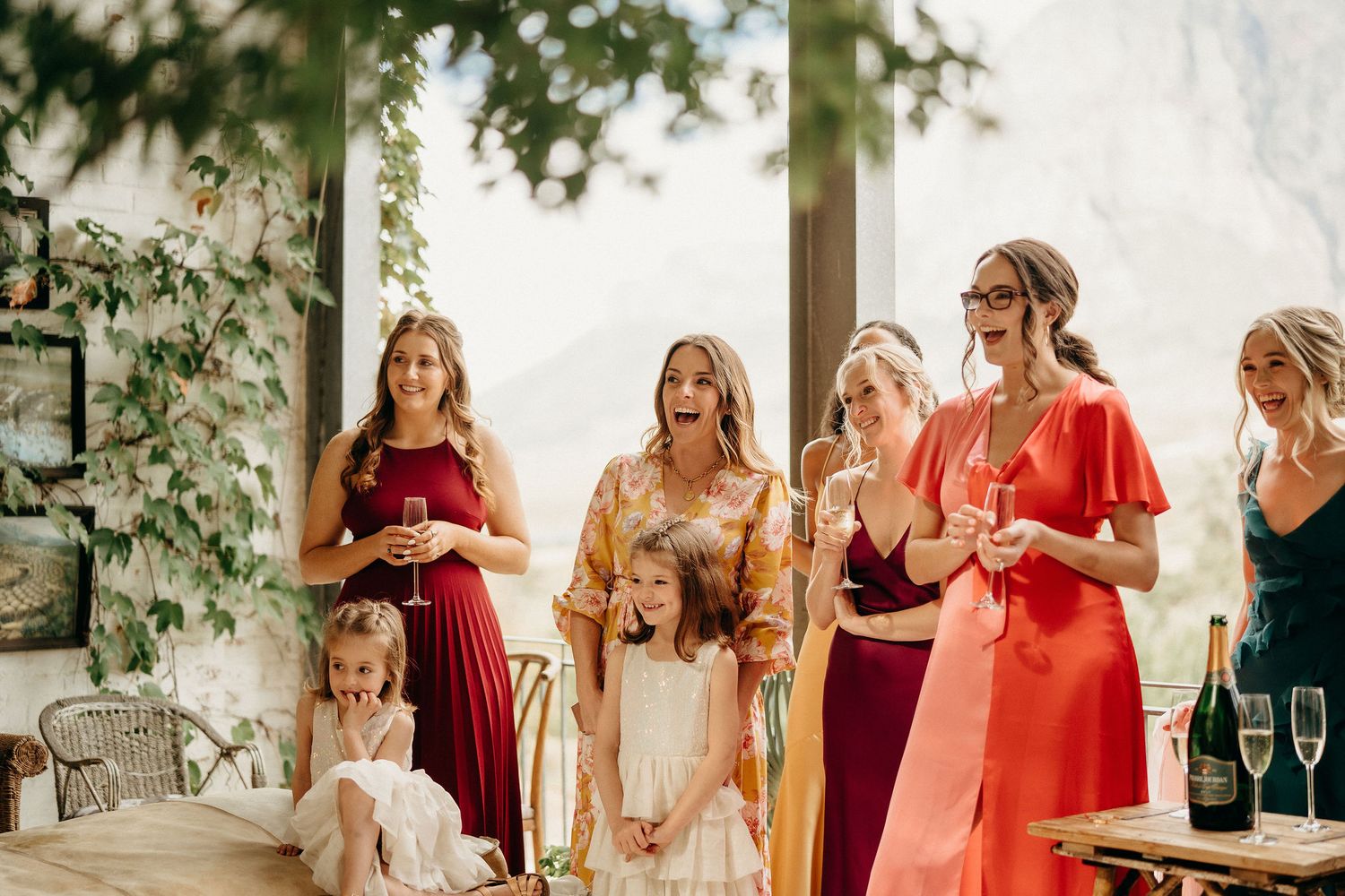 Wedding celebration with bridal party in vibrant red, yellow, and green dresses on an outdoor patio with champagne.