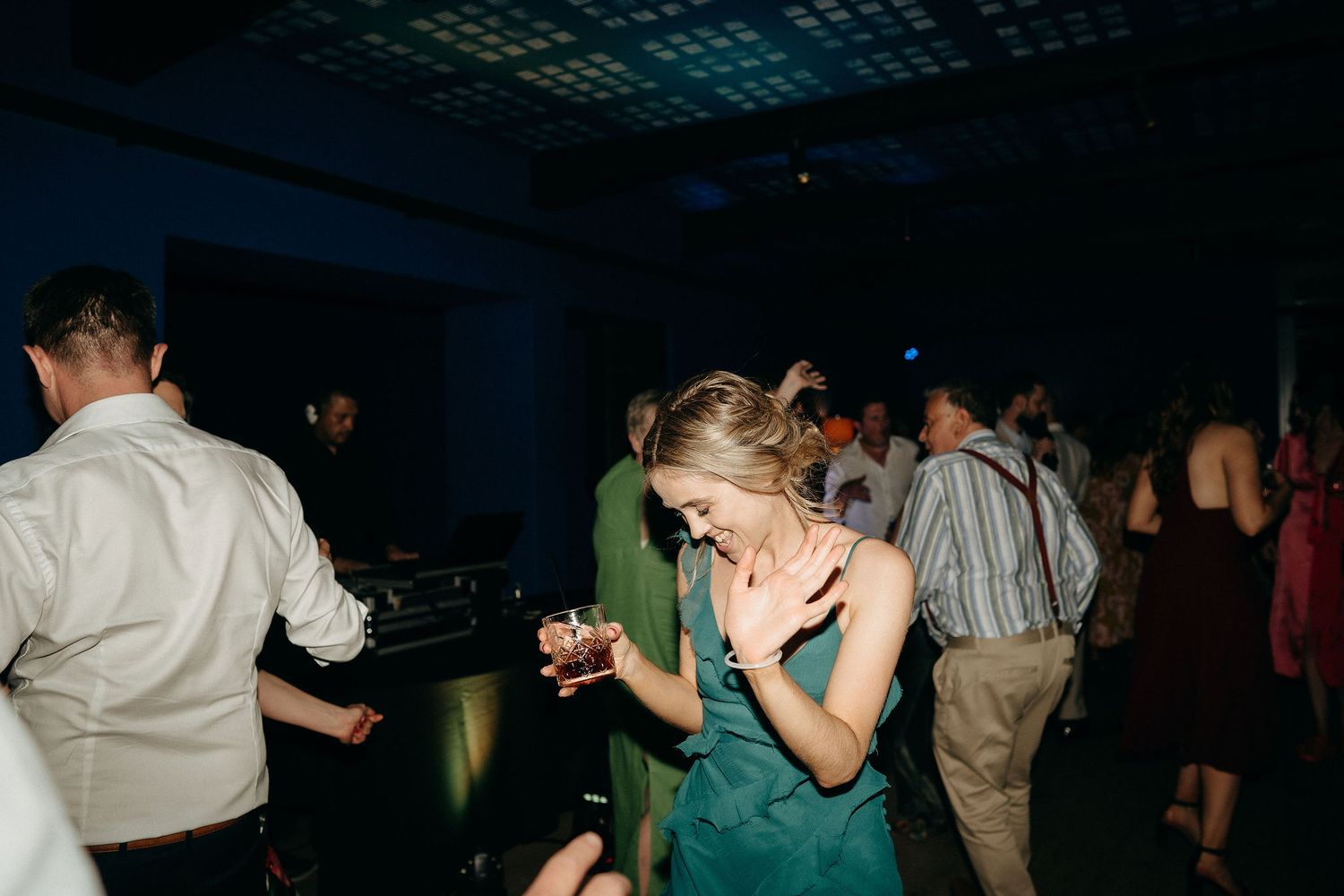 People dancing and mingling at a dimly lit party venue with someone in a green dress on the dance floor.