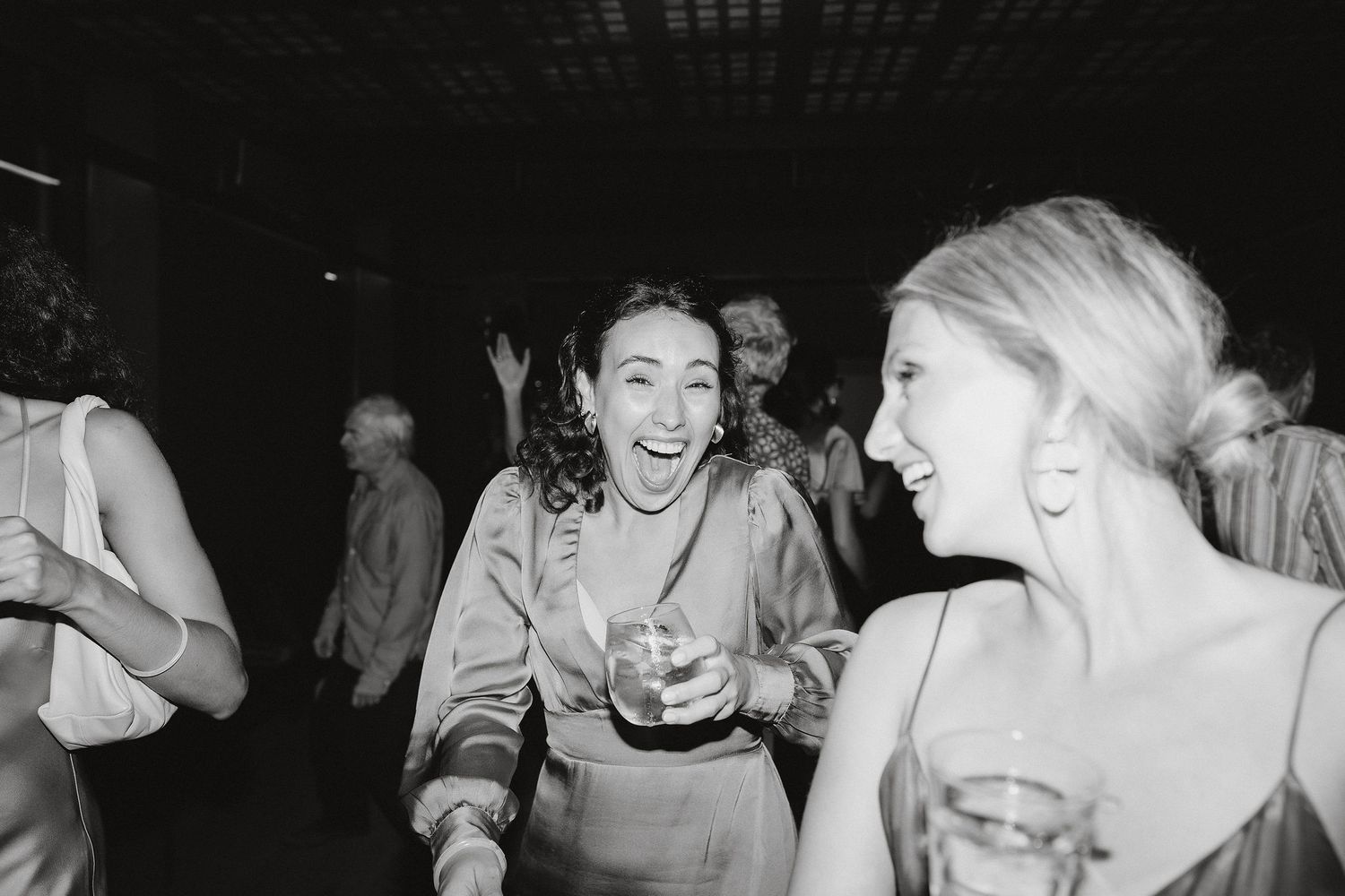 Black and white candid photo of two partygoers joyfully laughing while holding drinks at a social gathering.