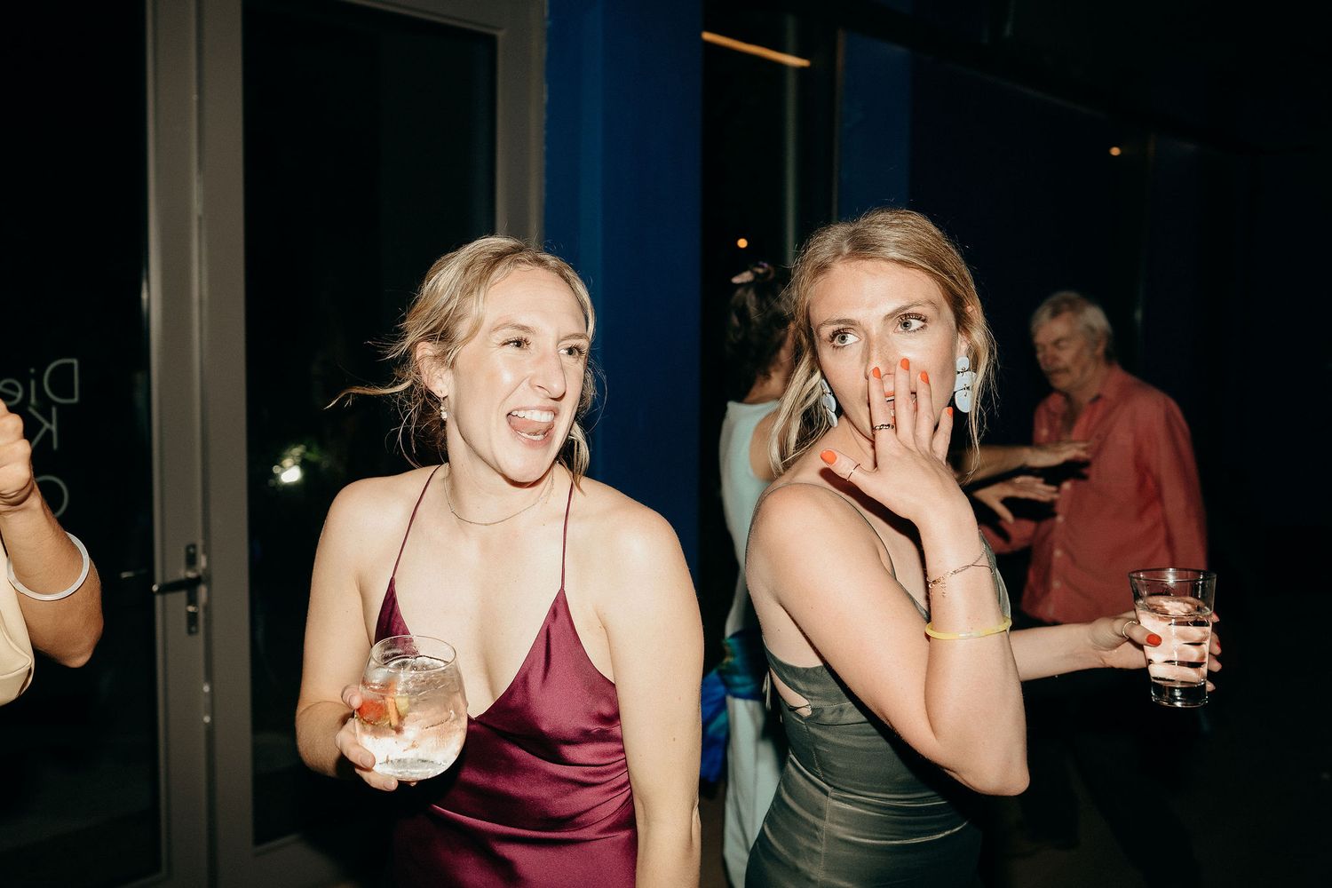 Two friends in formal dresses laugh and react with surprise while holding drinks at an evening event.