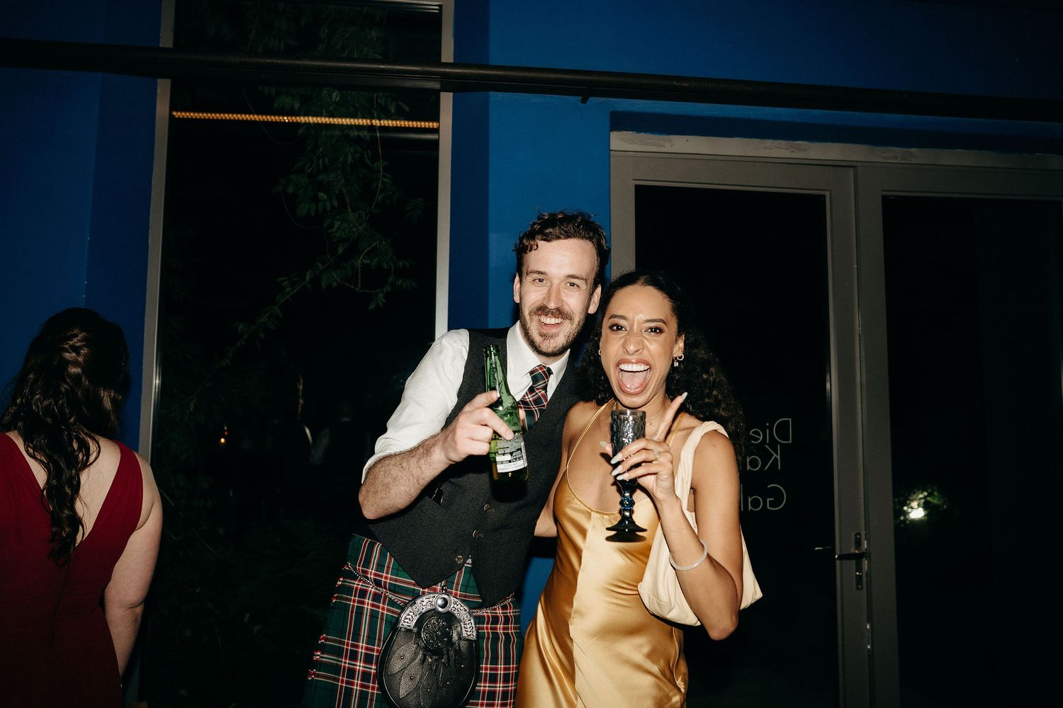 Party guests pose together during a wedding reception, wearing a traditional kilt and gold dress with drinks in hand.