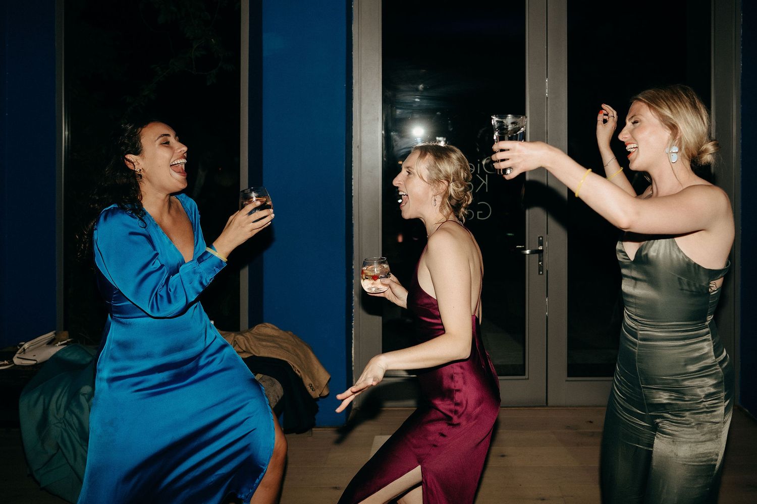 Three women in elegant dresses dancing and laughing together at a nighttime party.