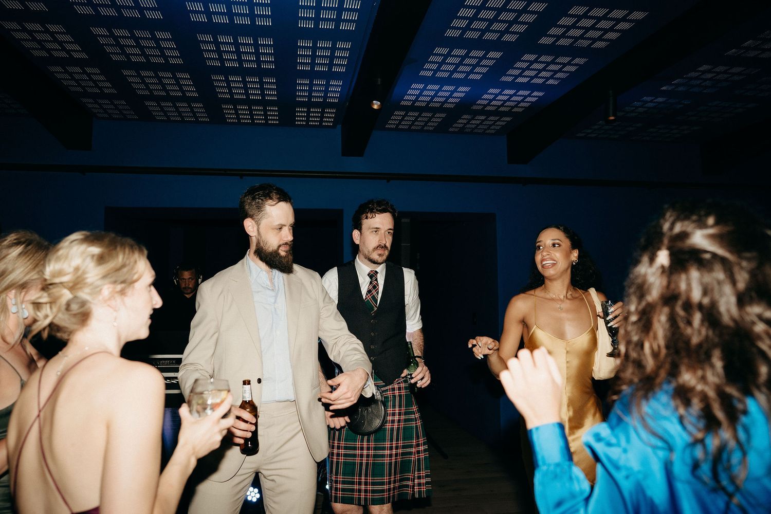 A candid moment at a wedding reception shows guests socializing and laughing on a dimly lit dance floor.
