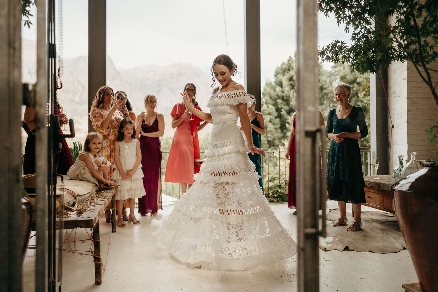 Wedding scene showing people gathered around a bride in a beautiful layered white gown against a scenic outdoor backdrop.
