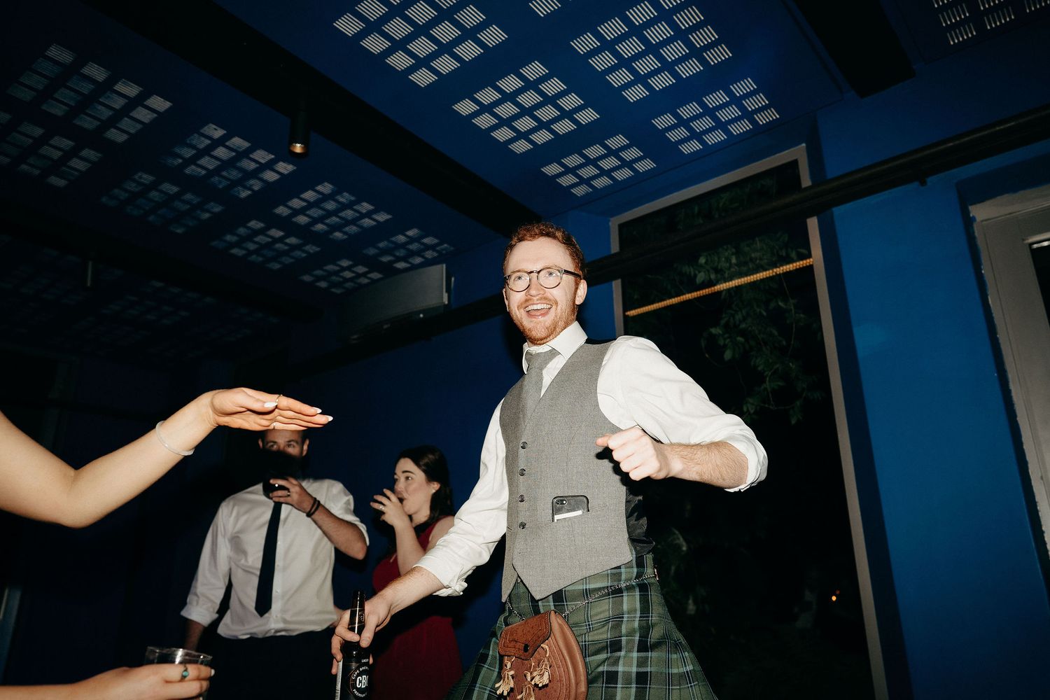 A person in vest and dress shirt dancing energetically at a party with a blue lit ceiling above.