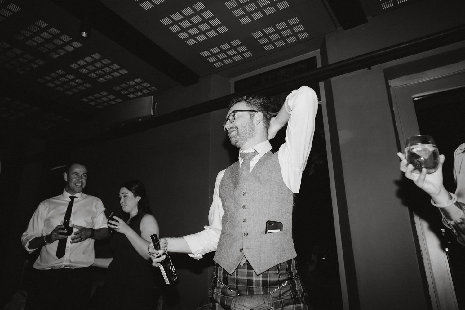 A person in a vest dances energetically at a party while others watch in the background of a dark room.