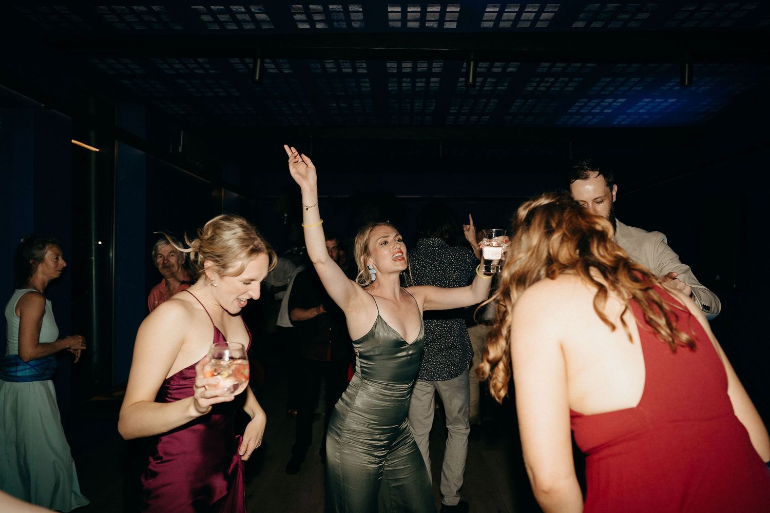 Wedding guests in red and green dresses dance excitedly together on a dimly lit dance floor.