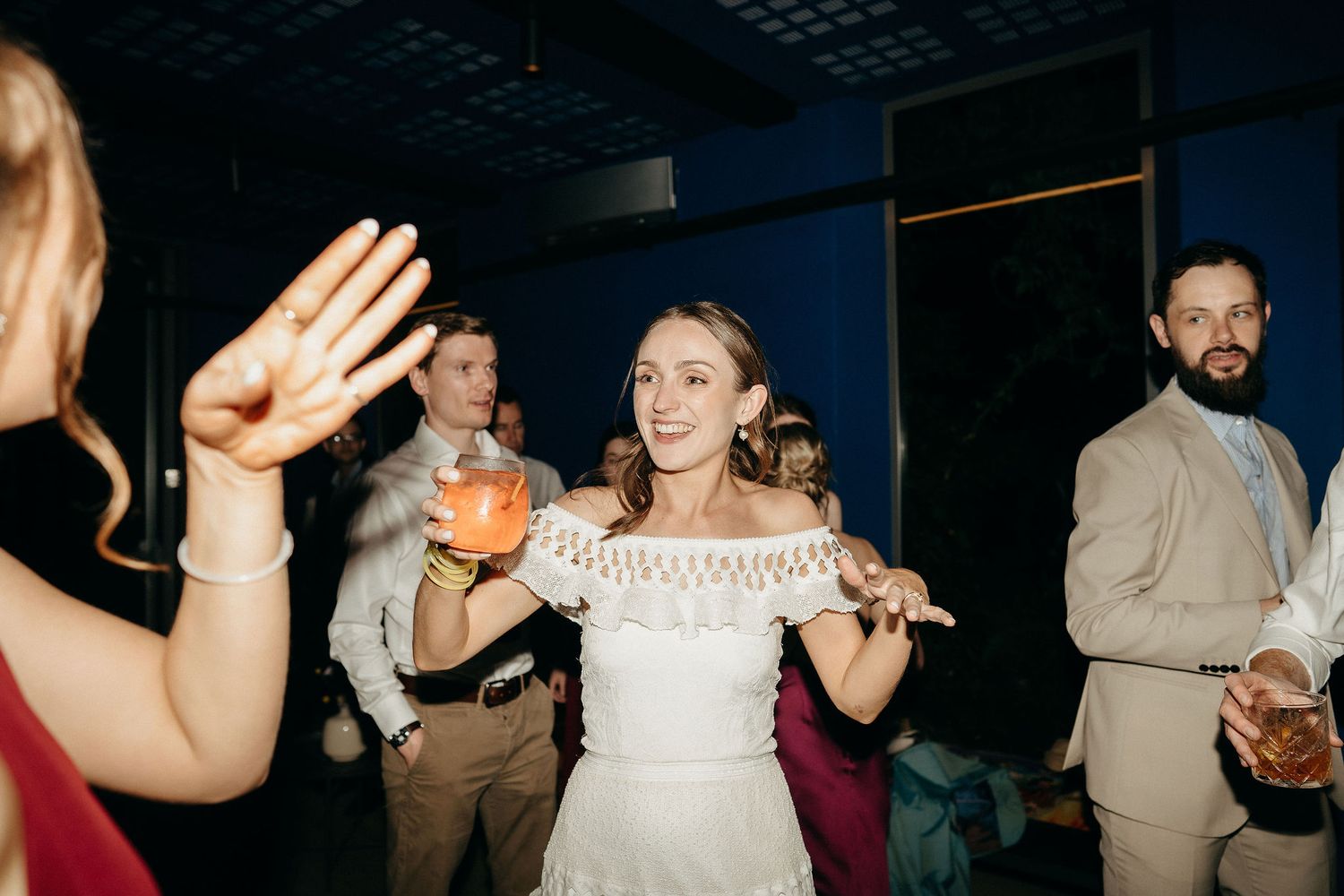 A lively wedding reception scene with a dancer in a white off-shoulder dress holding a drink while celebrating on the dance floor.