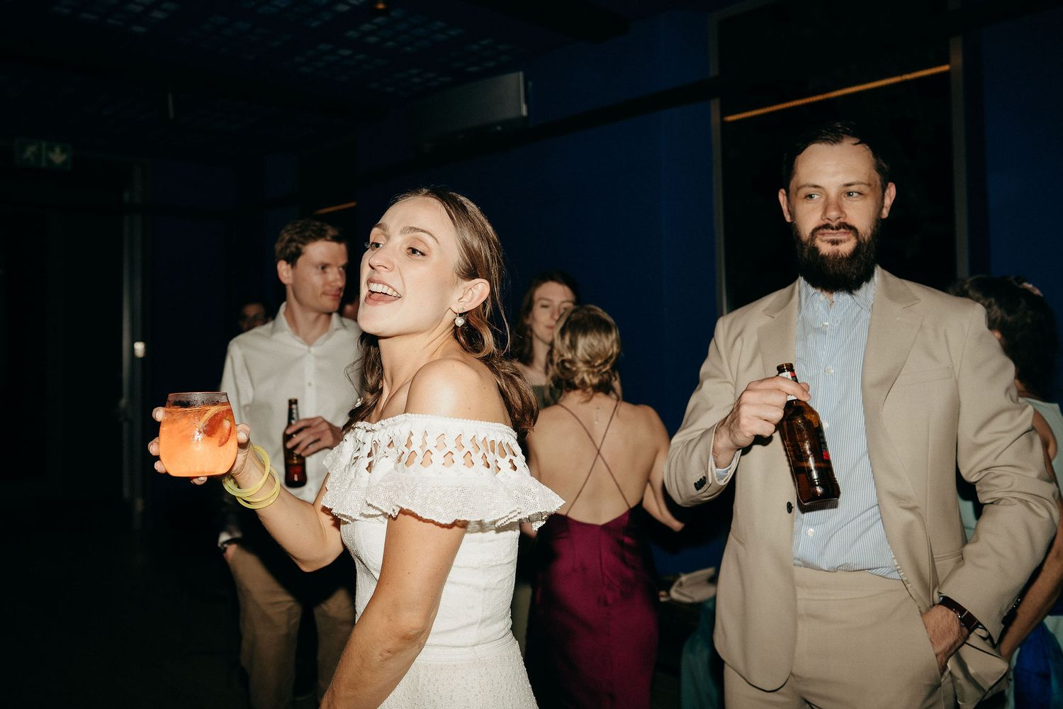 Partygoers enjoying drinks at a dimly lit evening celebration, with a person in white dress and another in beige suit.
