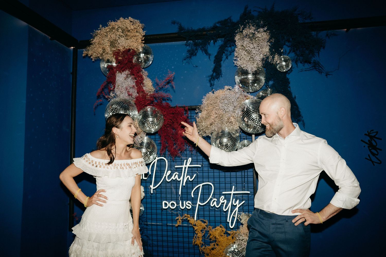 A couple poses together in front of a blue wall decorated with a neon sign saying Do the Party and shimmering decor.