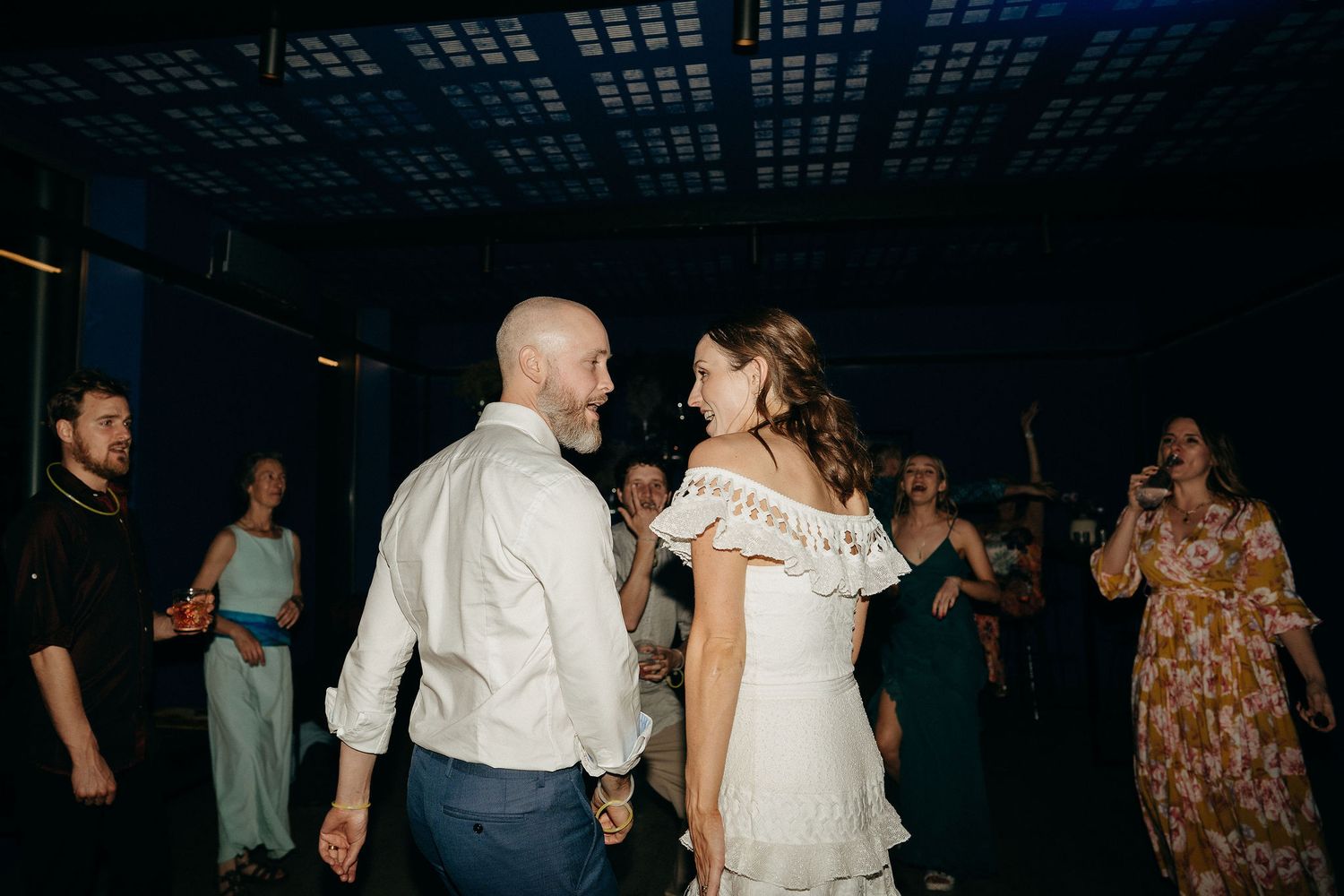 A couple dances together at a wedding reception while guests look on in a dimly lit venue.