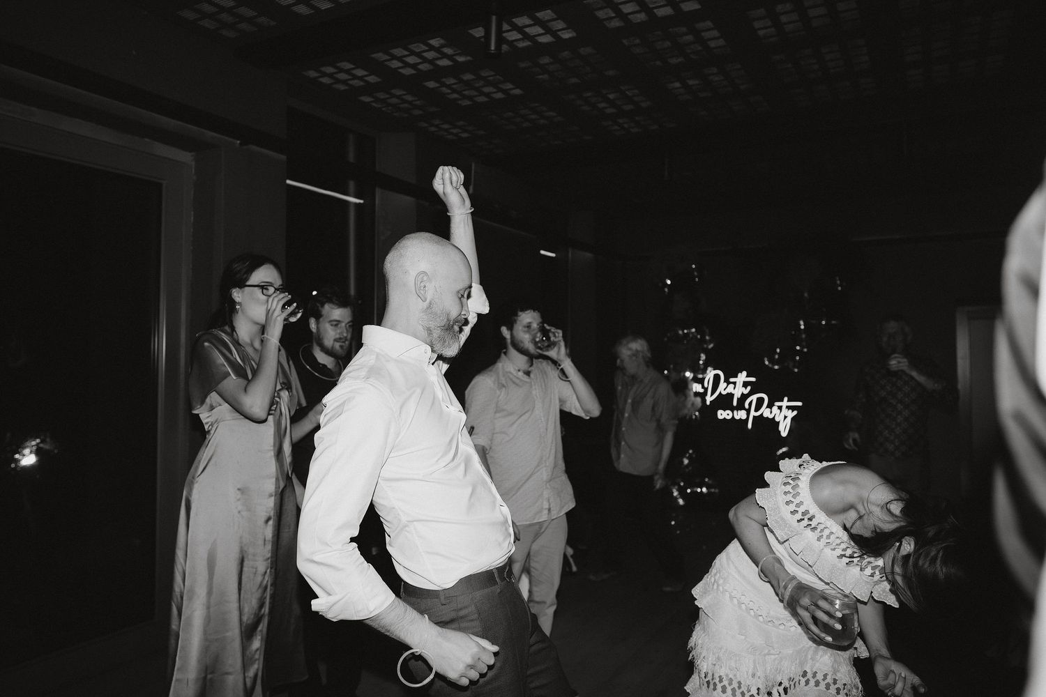 Black and white photo of wedding guests dancing and raising their arms on a darkened dance floor at a reception celebration.