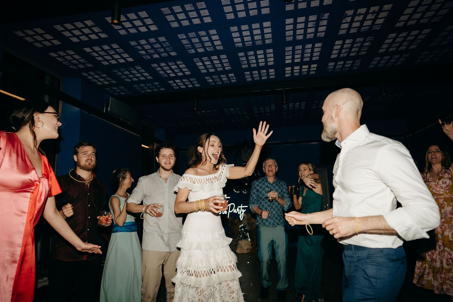 People dancing and having fun at a wedding reception under a dark ceiling with geometric lighting patterns.