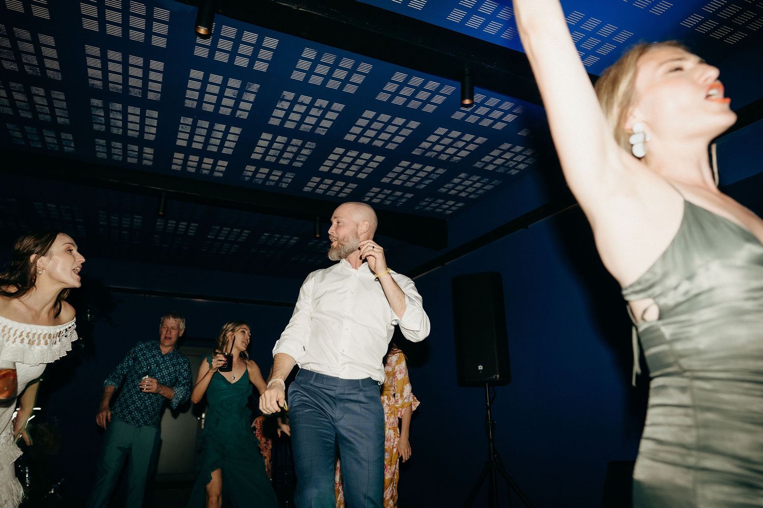 People dancing energetically at a reception under a dark ceiling with blue lighting.