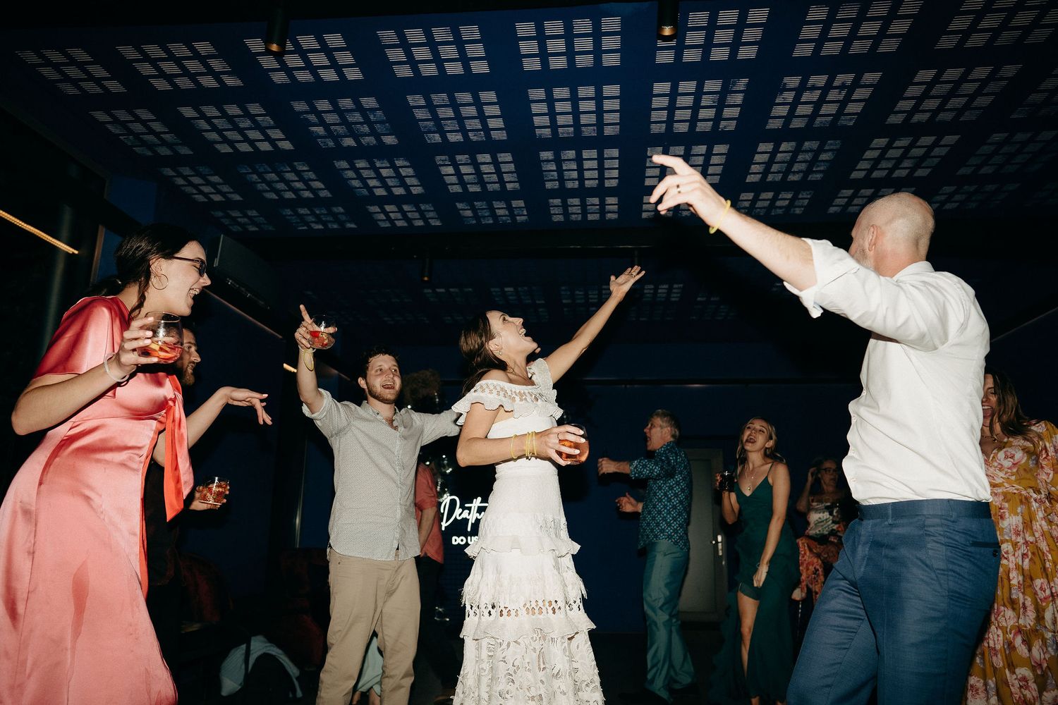 Wedding guests dancing and celebrating together under a dark ceiling with patterned lighting at a reception party.