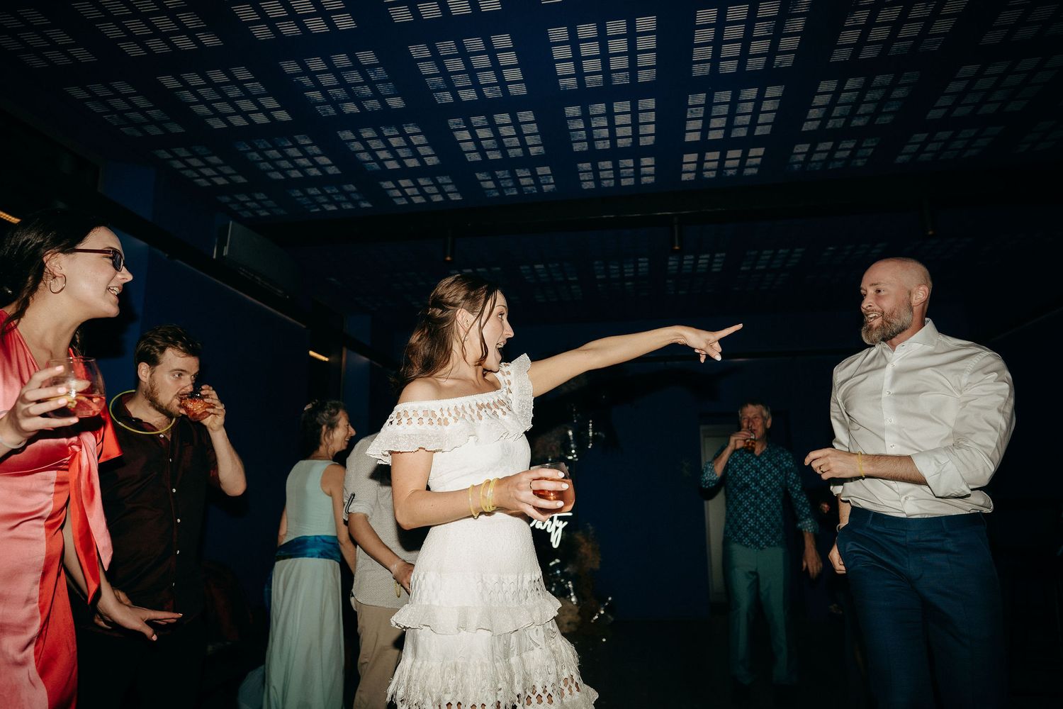 Joyful bride points while dancing with guests on a dimly lit dance floor during a wedding reception celebration.