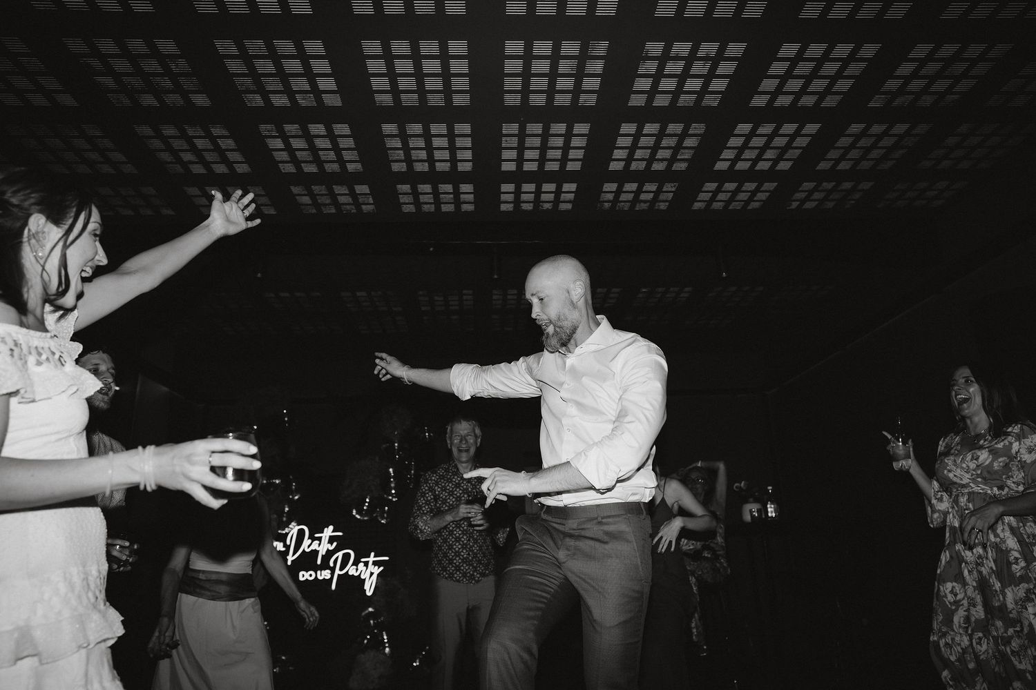 Black and white photo of energetic dancing at a reception under a modern skylight venue ceiling.