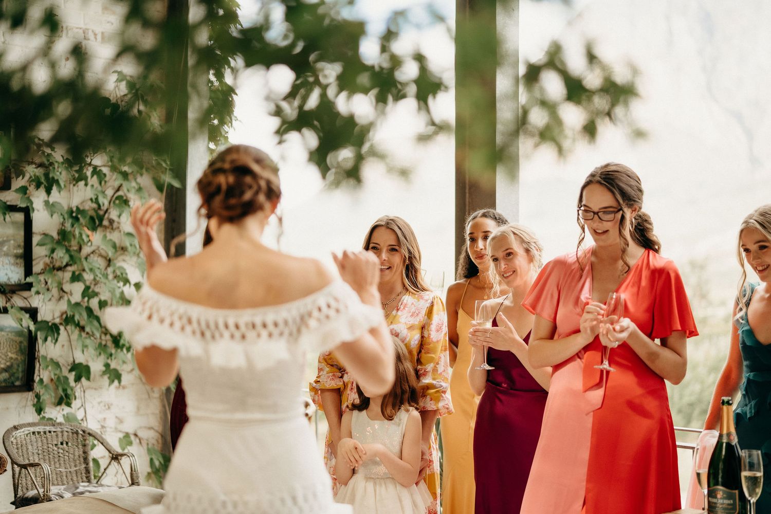 Group in colorful dresses at an outdoor wedding ceremony gathered under a covered area with greenery.
