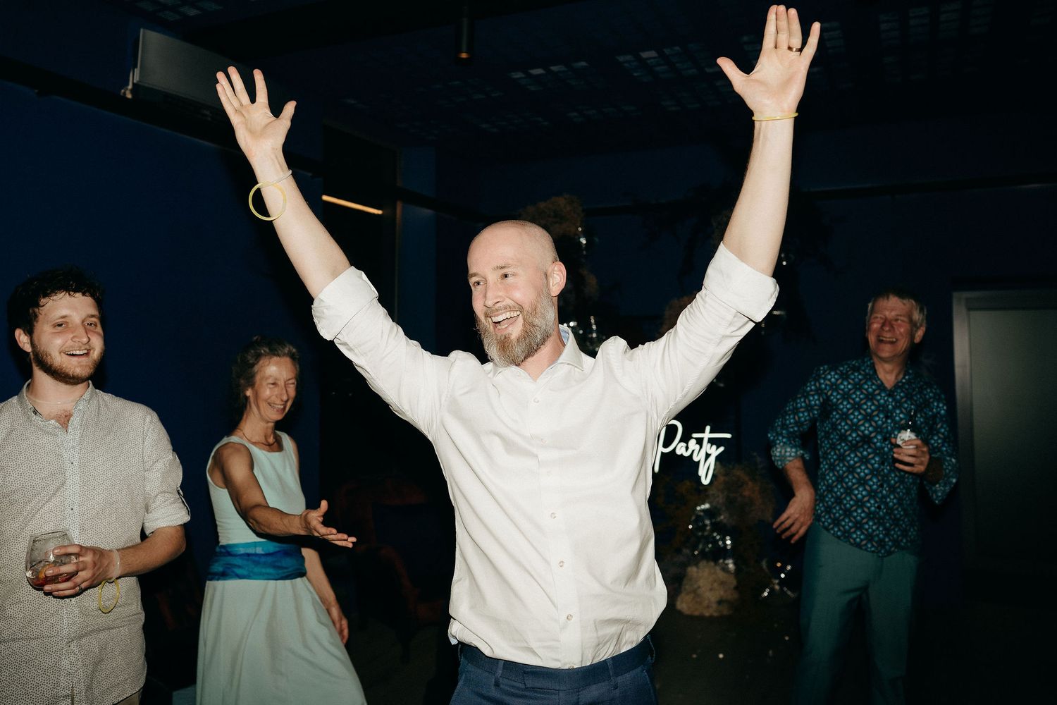 Guest in a white shirt raises their arms in celebration while dancing at a party with other guests.