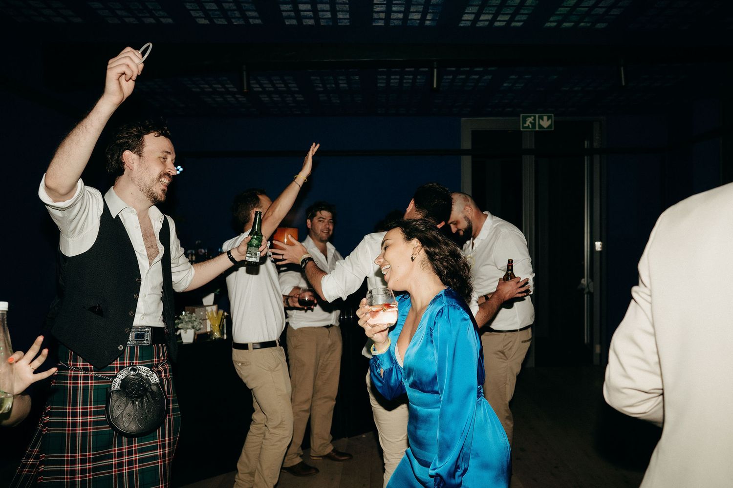 Wedding guests dance energetically on a dark dance floor while dressed in formal attire at a reception.