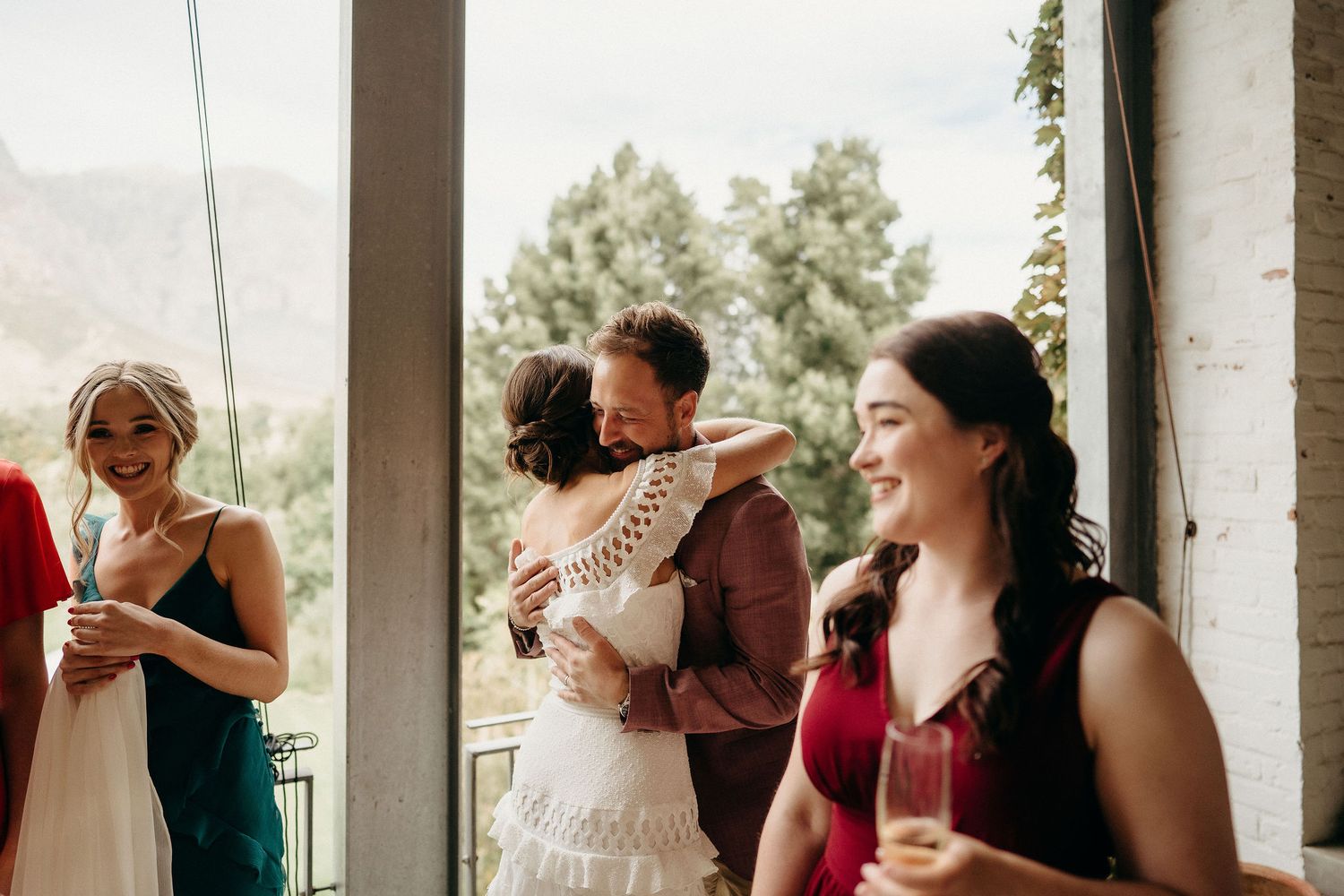People celebrate at an outdoor wedding reception with drinks and warm embraces under a covered porch area.