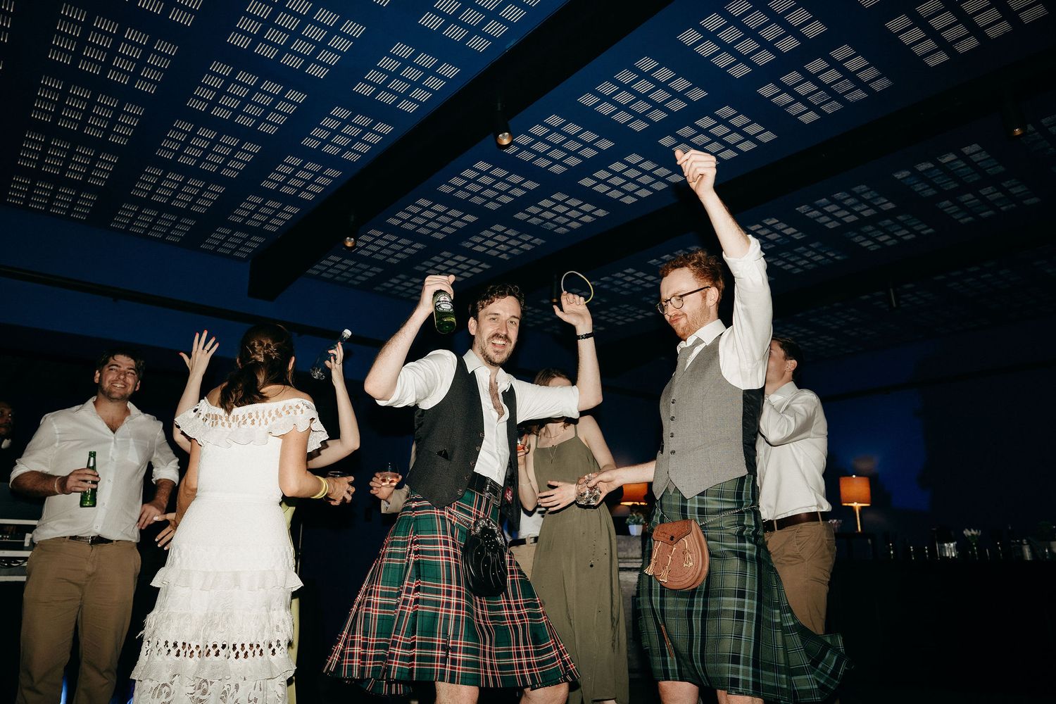 Wedding party guests in kilts and formal attire dance and celebrate with raised arms at an indoor reception.