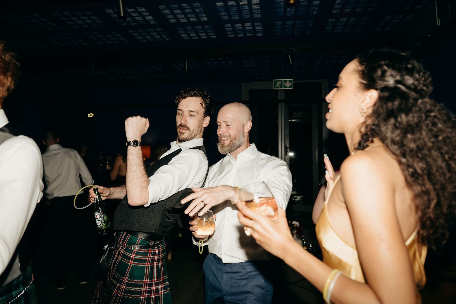 Party guests dance and celebrate at a wedding reception, with one person wearing a traditional Scottish kilt.