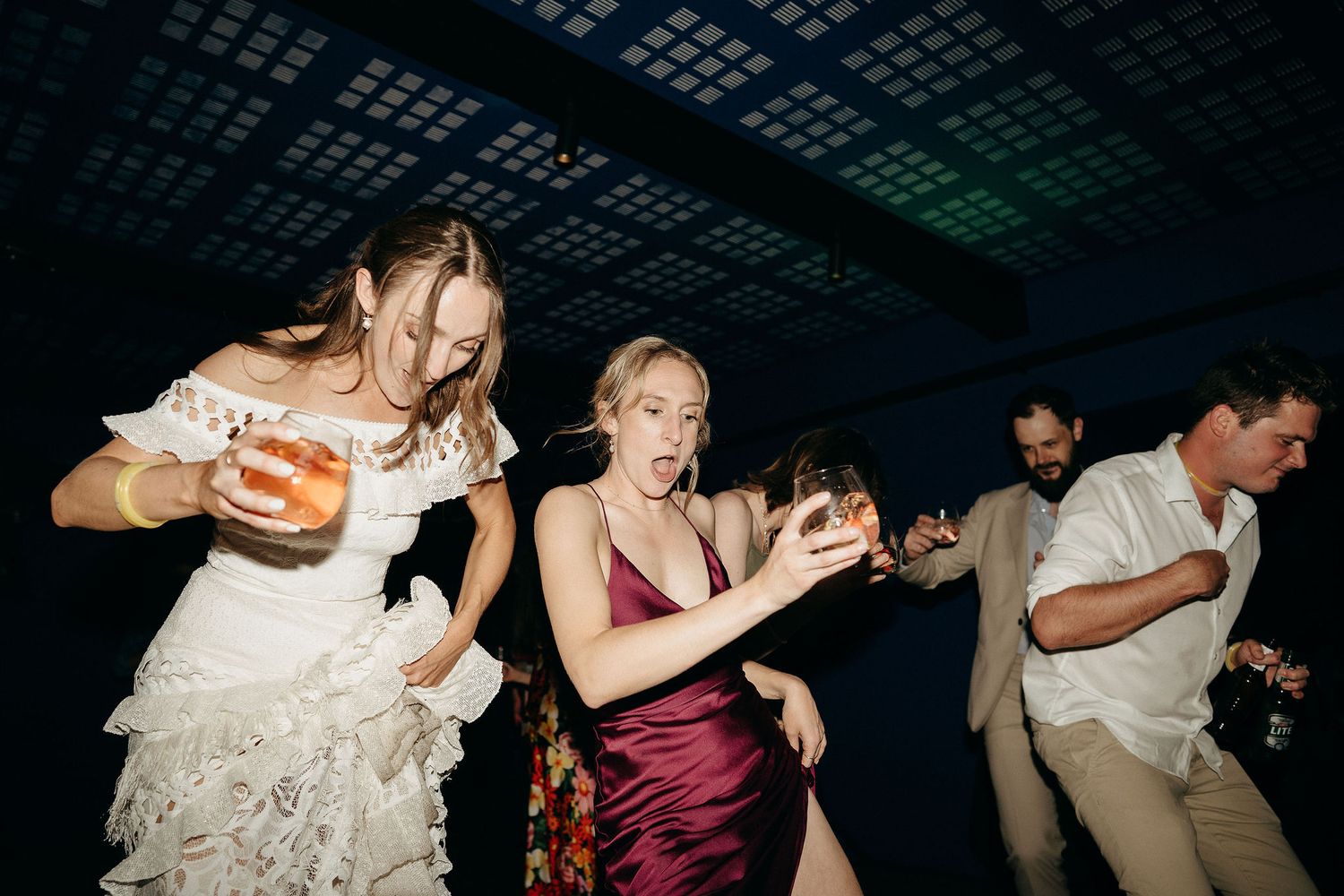 A group dances energetically on a dance floor beneath an ornate ceiling at a wedding reception.